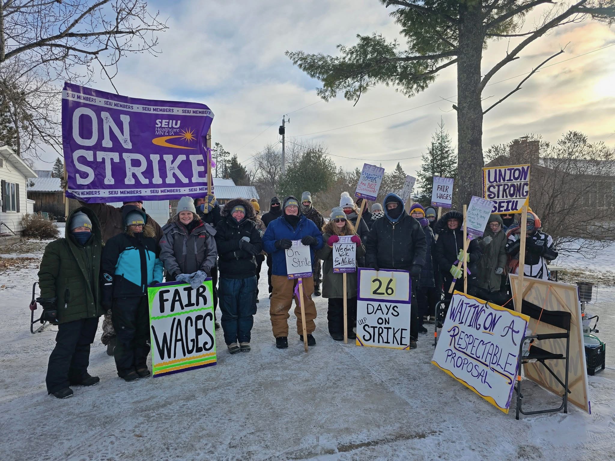 around 2 dozen people wearing winter gear, matching grey beanies, stand in the snow holding picket signs reading "on strike" and "fair wages" and "union strong SEIU"