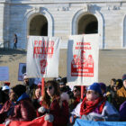 A crowd of people in winter gear stand in front of a while marble building, two hold signs with prints that say "proud union educator" and "strong schools strong communities" in red paint