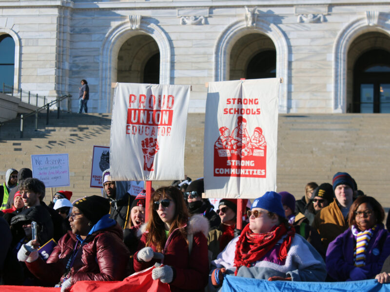 A crowd of people in winter gear stand in front of a while marble building, two hold signs with prints that say "proud union educator" and "strong schools strong communities" in red paint