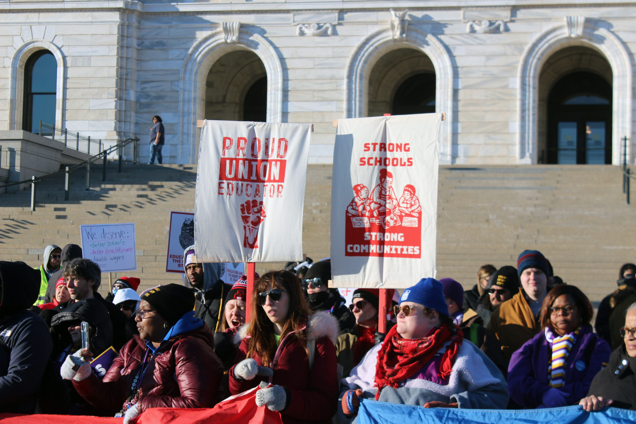 A crowd of people in winter gear stand in front of a while marble building, two hold signs with prints that say "proud union educator" and "strong schools strong communities" in red paint