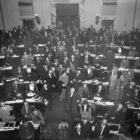 A black and white image showing rows of people standing at desks with books