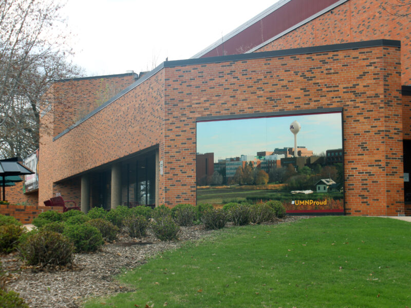 The brown brick exterior of the St. Paul campus food hall includes a photo of St. Paul campus behind green bushes and lawn