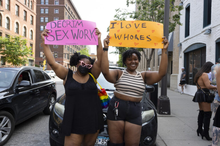 Two friends hold up signs, one reads, “Decrim sex work”, a central demand of SWOP MPLS, and the other reads, “I love whores”.