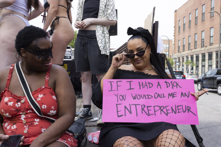 Two parade-goers pose for a photo with a sign that reads, “If I had a dick you would call me an entrepreneur”.