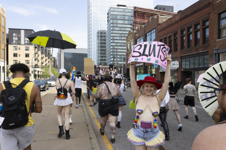 Marchers make their way down the street with a smiling attendee with a sign that reads “Sluts” and a bible tucked into their shorts.