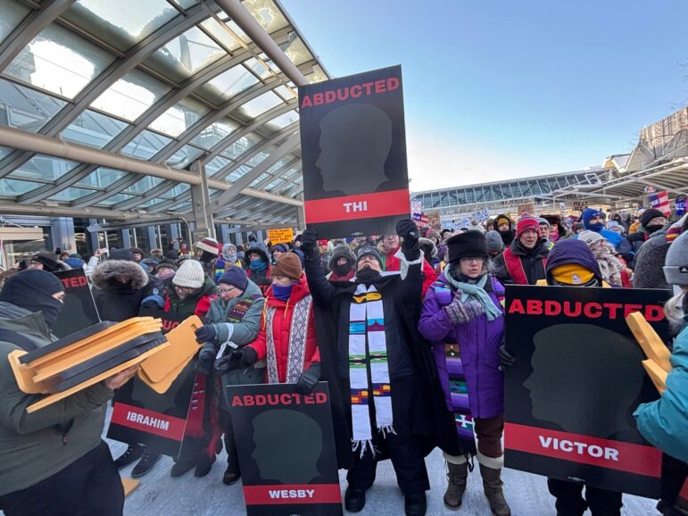 A crowd of people dressed in winter gear and holding signs with the names of people abducted by ICE walk outside near the airport terminal